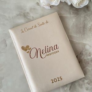 May include: A cream-colored notebook with the text "Le Carnet de Sant&eacute; de" at the top. The name "Melina Rastoder" is written in a glittery font, with the year "2025" below. A bouquet of white roses is in the background.