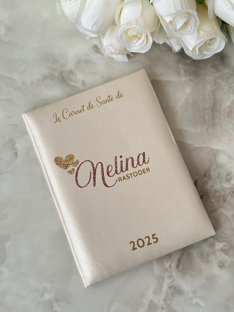 May include: A cream-colored notebook with the text "Le Carnet de Sant&eacute; de" at the top. The name "Melina Rastoder" is written in a glittery font, with the year "2025" below. A bouquet of white roses is in the background.
