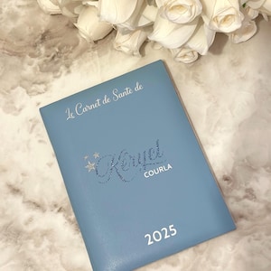 May include: A light blue notebook with the text "Le Carnet de Sant&eacute; de Keryel COURLA 2025" in silver lettering. White roses are in the background.