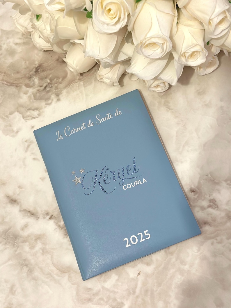 May include: A light blue notebook with the text "Le Carnet de Sant&eacute; de Keryel COURLA 2025" in silver lettering. White roses are in the background.