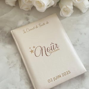May include: Cream-colored journal with gold lettering and the text "Le Carnet de Sant&eacute; de Noir Qulbani 02 JUIN 2025". The journal is closed and has a soft, leather-like texture. White roses are in the background.