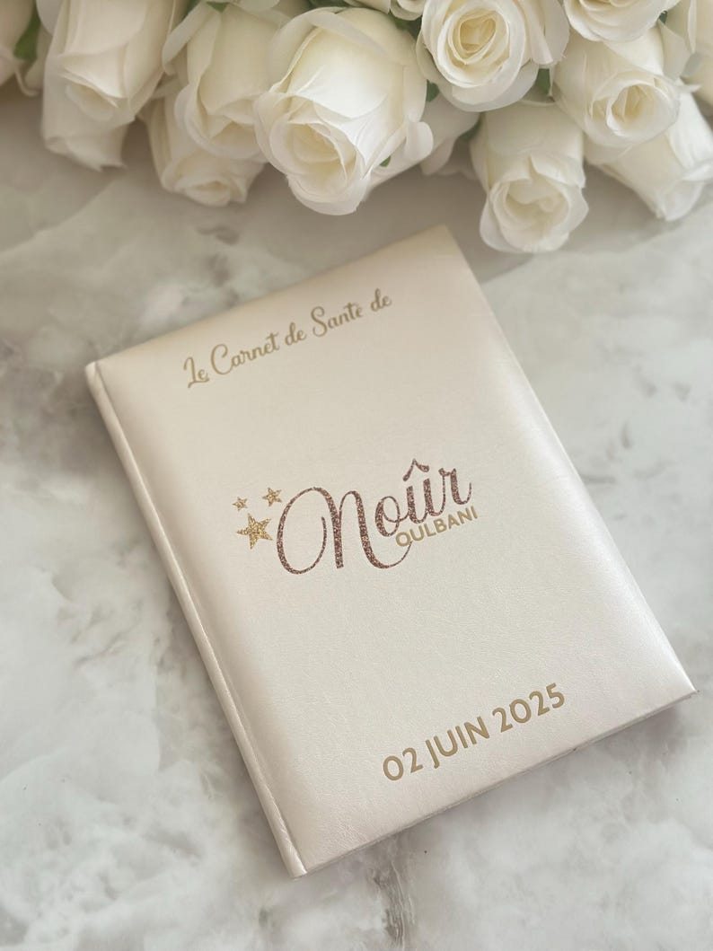 May include: Cream-colored journal with gold lettering and the text "Le Carnet de Sant&eacute; de Noir Qulbani 02 JUIN 2025". The journal is closed and has a soft, leather-like texture. White roses are in the background.