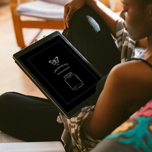 Puede incluir: Una tableta negra muestra un dibujo lineal blanco de una mariposa y un tarro, con el texto "The Limiting Beliefs Release Workbook". La tableta la sostiene una persona sentada en un sofá, con un libro en su regazo.