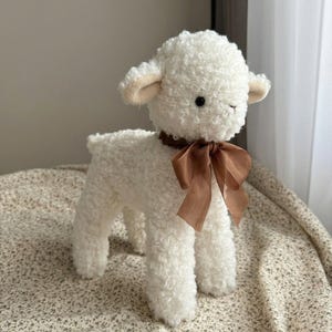 Puede incluir: Un juguete de peluche de cordero blanco con textura rizada. El cordero tiene una cinta marr&oacute;n atada al cuello y peque&ntilde;as orejas beige. El juguete est&aacute; sobre una tela estampada, con un fondo de color neutro.