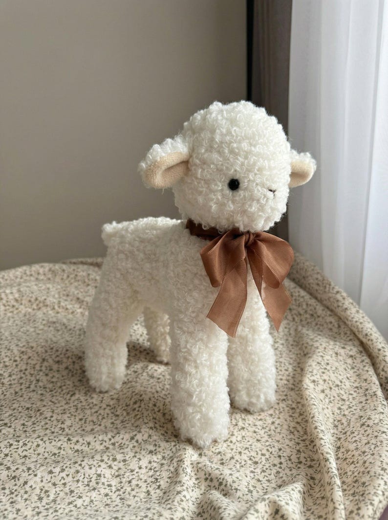 Puede incluir: Un juguete de peluche de cordero blanco con textura rizada. El cordero tiene una cinta marr&oacute;n atada al cuello y peque&ntilde;as orejas beige. El juguete est&aacute; sobre una tela estampada, con un fondo de color neutro.