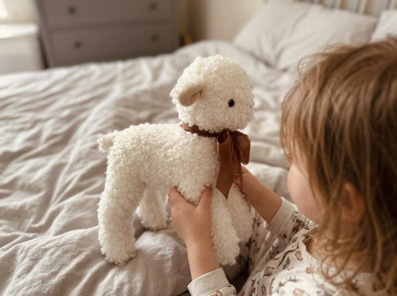 Puede incluir: Un juguete de peluche de cordero blanco con una cinta marr&oacute;n alrededor del cuello. El cordero tiene una textura rizada y orejas peque&ntilde;as y redondeadas. El juguete es sostenido por una persona, lo que sugiere un art&iacute;culo para ni&ntilde;os.