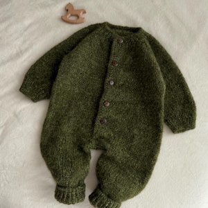 May include: A green knitted baby romper with long sleeves and button fastenings. The romper is made of a soft, textured yarn and features ribbed cuffs and ankles. A small wooden rocking horse toy is next to the romper.