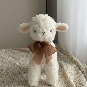Puede incluir: Un peluche de cordero blanco con una textura rizada. El cordero tiene ojos peque&ntilde;os y oscuros, orejas beige y una cinta marr&oacute;n atada alrededor del cuello. El juguete est&aacute; sobre una tela con estampado floral.