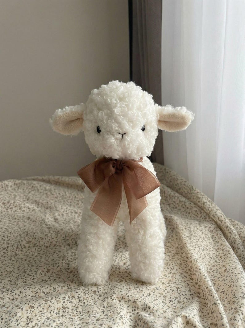 Puede incluir: Un peluche de cordero blanco con una textura rizada. El cordero tiene ojos peque&ntilde;os y oscuros, orejas beige y una cinta marr&oacute;n atada alrededor del cuello. El juguete est&aacute; sobre una tela con estampado floral.