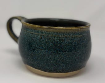 May include: A handmade ceramic mug with a dark teal glaze and a tan base. The mug has a rounded shape with a handle and a slightly flared rim. The glaze has a speckled appearance, adding texture to the piece.