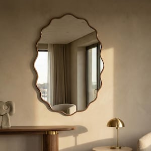 May include: An interior scene with a large, wavy-edged mirror in a bronze frame. A wooden console table with a decorative sculpture is positioned below the mirror. A small side table with a brass lamp and a cream armchair are also present.