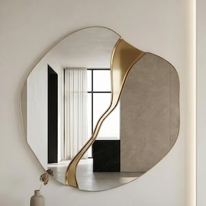 May include: An irregularly shaped wall mirror with a gold-colored frame and a gold accent. The mirror is mounted on a white wall above a black rectangular base. A small vase with dried flowers sits on the base.