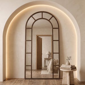 May include: A tall, arched floor mirror with a dark brown metal frame divided into rectangular panes. The mirror features a rounded top and a grid design, reflecting a room with a wooden door, a bed, and a lamp. It stands on a wooden floor.