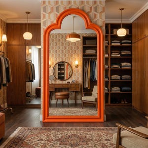 May include: A stylish walk-in closet with a large orange framed mirror. The room features wooden cabinets, a clothing rack, and a patterned rug. The walls are decorated with a geometric wallpaper, and the room is illuminated by warm lighting.