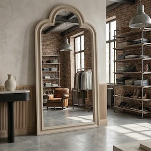 May include: A large, arched mirror with a light-colored frame dominates the room. The mirror reflects a brick wall, shelving units with folded clothes, and a clothing rack. A leather armchair and industrial-style lighting complete the interior design.