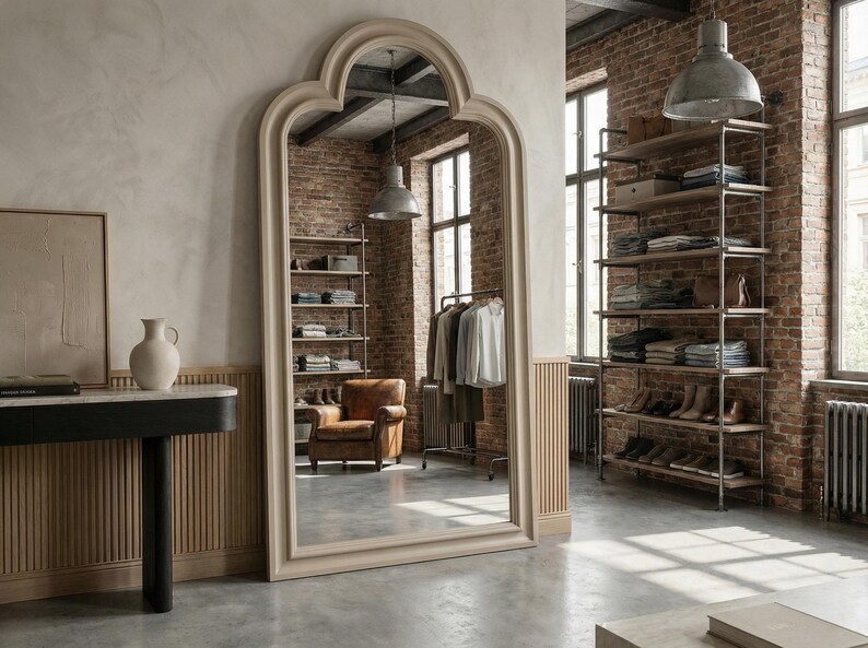 May include: A large, arched mirror with a light-colored frame dominates the room. The mirror reflects a brick wall, shelving units with folded clothes, and a clothing rack. A leather armchair and industrial-style lighting complete the interior design.