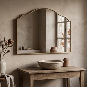 May include: A wall-mounted mirror with a thin gold-toned frame featuring a curved, arched top edge. The mirror reflects a room with a window, a small bottle, and a terracotta-coloured jar. The frame has a smooth, metallic finish and a clean design.