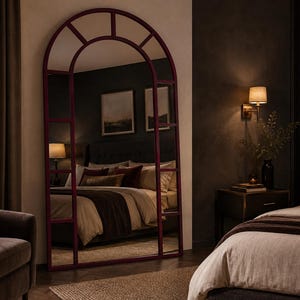 May include: A tall, floor-standing arched mirror with a deep burgundy frame. The mirror features a grid-like windowpane design with multiple rectangular sections and a semi-circular top. The reflective surface shows a bedroom interior with a bed.