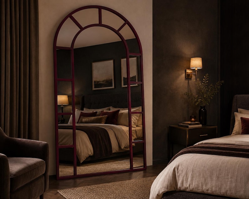 May include: A tall, floor-standing arched mirror with a deep burgundy frame. The mirror features a grid-like windowpane design with multiple rectangular sections and a semi-circular top. The reflective surface shows a bedroom interior with a bed.