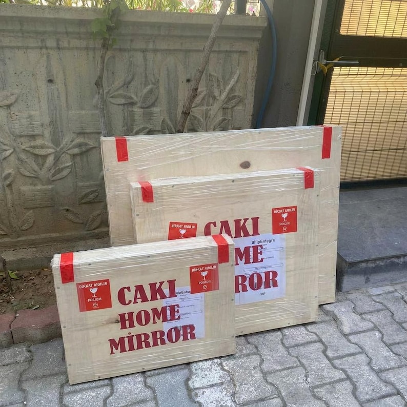 May include: Three rectangular wooden shipping crates of varying sizes, wrapped in clear plastic and secured with red tape. Each crate features a white label with the red text CAKI HOME MIRROR and a red fragile symbol sticker on the front.