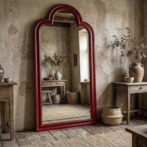 May include: A large, ornate, red-framed mirror leans against a textured wall. The mirror reflects a rustic interior with wooden furniture, vases, and woven baskets. The room has a warm, inviting atmosphere with natural light.
