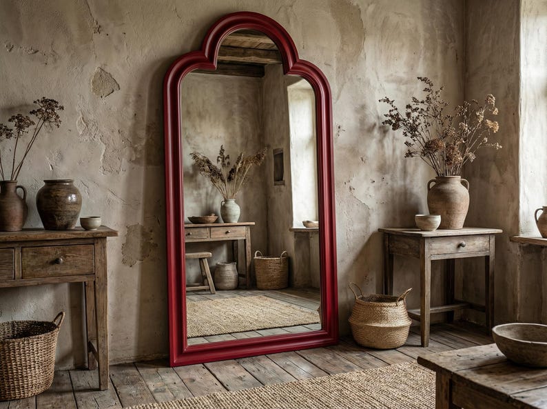 May include: A large, ornate, red-framed mirror leans against a textured wall. The mirror reflects a rustic interior with wooden furniture, vases, and woven baskets. The room has a warm, inviting atmosphere with natural light.