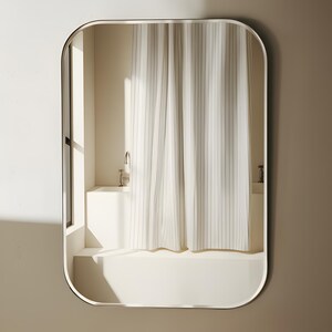 May include: A rectangular mirror with rounded corners, framed in a thin, dark metal. The mirror reflects a bathroom scene with a white bathtub, a shower curtain with vertical stripes, and a window.