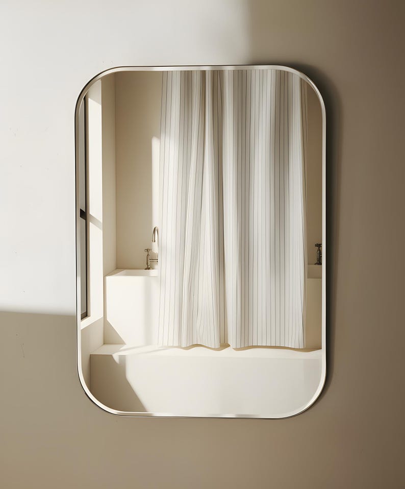 May include: A rectangular mirror with rounded corners, framed in a thin, dark metal. The mirror reflects a bathroom scene with a white bathtub, a shower curtain with vertical stripes, and a window.