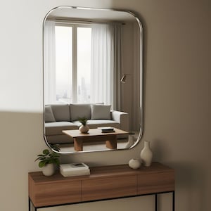 May include: A rectangular mirror with curved edges is mounted above a wooden console table. The mirror reflects a living room scene with a grey sofa, coffee table, and window. The console holds vases, a potted plant, and books.