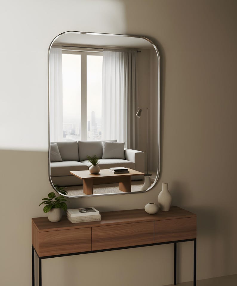May include: A rectangular mirror with curved edges is mounted above a wooden console table. The mirror reflects a living room scene with a grey sofa, coffee table, and window. The console holds vases, a potted plant, and books.