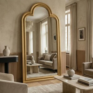 May include: A large, gold-framed mirror with an arched top stands in a neutral-toned room. The mirror reflects a living room with a sofa, coffee table, and decorative vases. The room has a warm, inviting atmosphere.