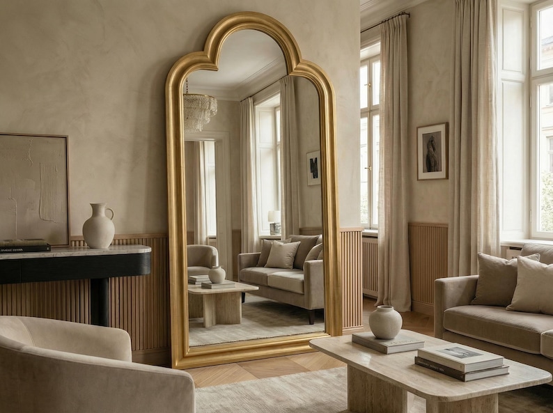 May include: A large, gold-framed mirror with an arched top stands in a neutral-toned room. The mirror reflects a living room with a sofa, coffee table, and decorative vases. The room has a warm, inviting atmosphere.