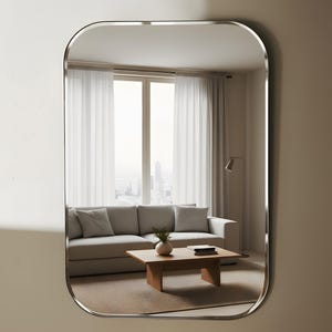 May include: A rectangular mirror with curved edges and a silver frame. The mirror reflects a living room scene with a grey sofa, wooden coffee table, and sheer white curtains. The room has a neutral colour palette.