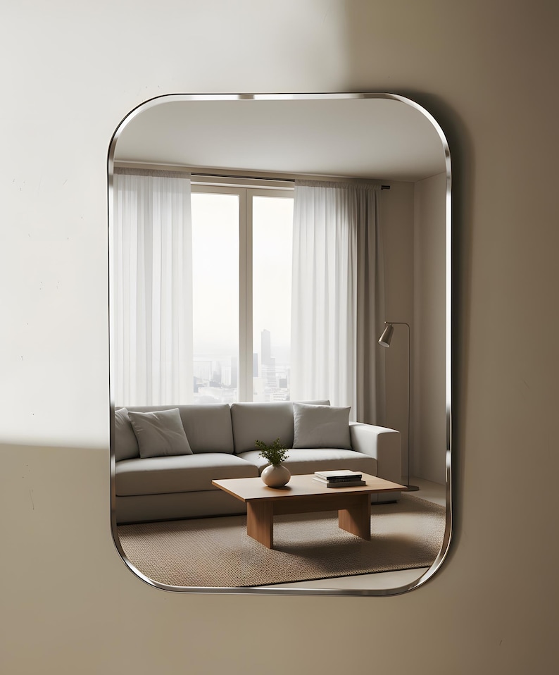 May include: A rectangular mirror with curved edges and a silver frame. The mirror reflects a living room scene with a grey sofa, wooden coffee table, and sheer white curtains. The room has a neutral colour palette.