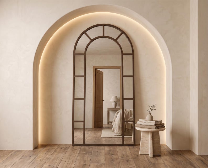 May include: A tall, arched floor mirror with a dark brown metal frame divided into rectangular panes. The mirror features a rounded top and a grid design, reflecting a room with a wooden door, a bed, and a lamp. It stands on a wooden floor.