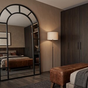 May include: A tall, floor-standing arched mirror with a black metal frame divided into rectangular panes. The mirror reflects a bedroom interior featuring a brown leather bench, a bed with neutral bedding, and dark wood cabinetry.
