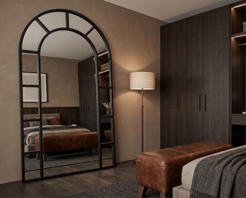 May include: A tall, floor-standing arched mirror with a black metal frame divided into rectangular panes. The mirror reflects a bedroom interior featuring a brown leather bench, a bed with neutral bedding, and dark wood cabinetry.