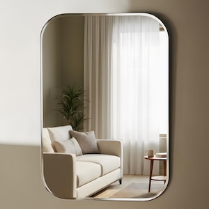 May include: A rectangular mirror with curved corners and a silver frame. The mirror reflects a living room scene with a cream sofa, cushions, a small table, and a plant. The room has neutral tones and natural light.