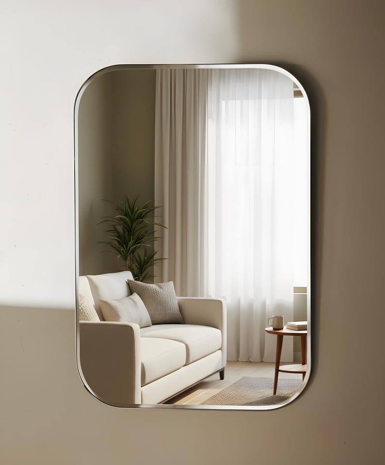May include: A rectangular mirror with curved corners and a silver frame. The mirror reflects a living room scene with a cream sofa, cushions, a small table, and a plant. The room has neutral tones and natural light.