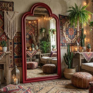 May include: A bohemian-style room with a large red-framed mirror, patterned rugs, and hanging plants. The room features a mix of textures, including woven textiles, leather poufs, and wooden furniture, creating a warm and inviting atmosphere. Candles and string lights add to the cozy ambiance.