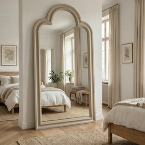 May include: A large, arched-top floor mirror with a light beige frame stands in a bright bedroom. The mirror reflects a bed with white linens, a wooden nightstand, and a framed botanical print. The room has neutral tones and natural light.