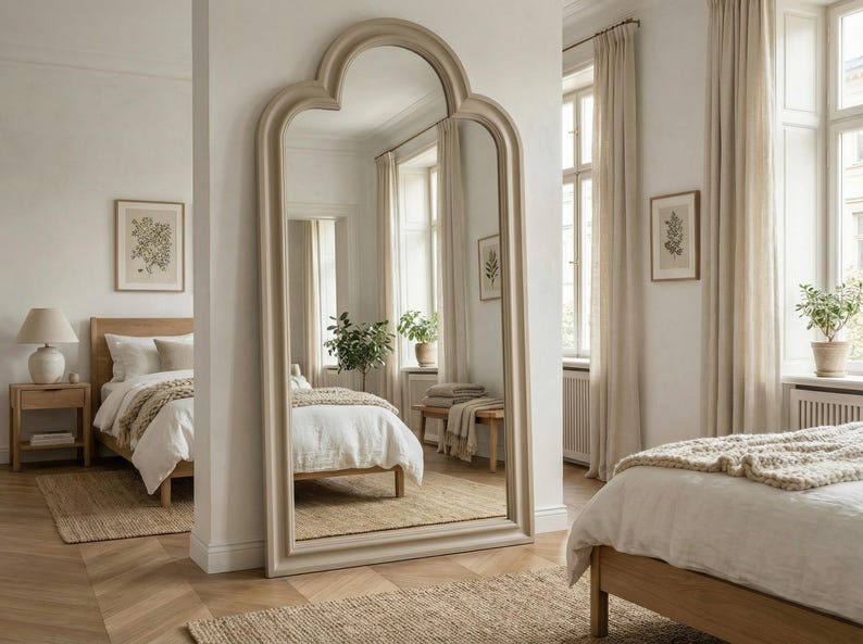 May include: A large, arched-top floor mirror with a light beige frame stands in a bright bedroom. The mirror reflects a bed with white linens, a wooden nightstand, and a framed botanical print. The room has neutral tones and natural light.