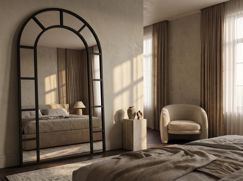 May include: A large, floor-standing arched mirror with a black metal frame divided into rectangular panes. The mirror features a curved top section and a grid-like structure, reflecting a bedroom interior with a bed, bedding, and a lamp.