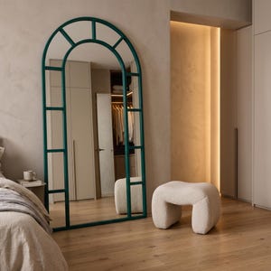 May include: A tall, floor-standing arched mirror with a dark teal metal frame divided into rectangular panes. The mirror features a rounded top section and a straight base, reflecting the surrounding room and a small, cream-colored textured stool.