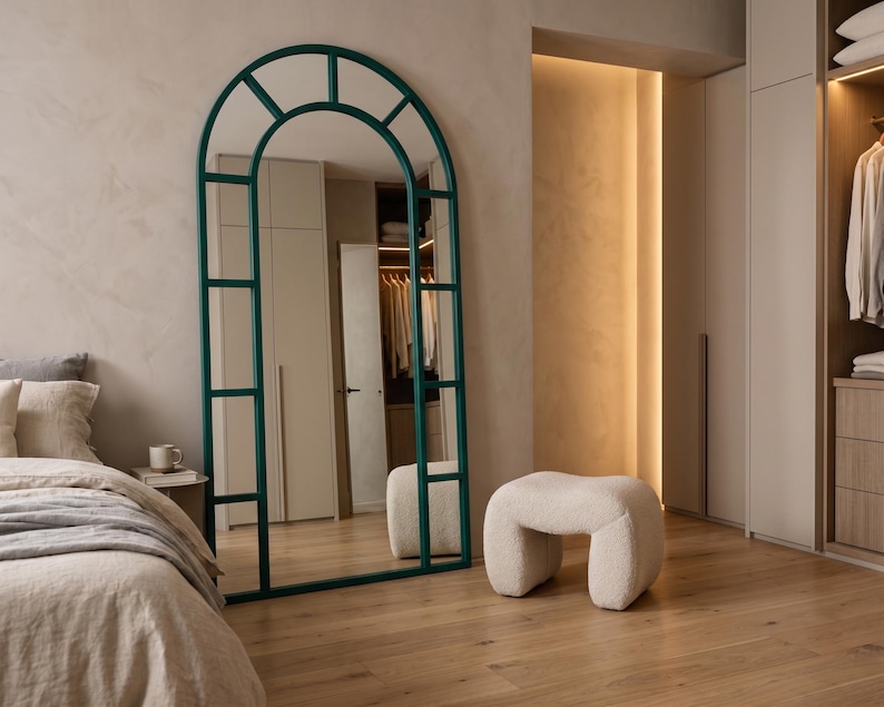May include: A tall, floor-standing arched mirror with a dark teal metal frame divided into rectangular panes. The mirror features a rounded top section and a straight base, reflecting the surrounding room and a small, cream-colored textured stool.