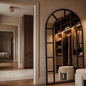 May include: A tall, arched floor mirror with a black metal frame featuring a grid design. The mirror reflects a walk-in closet with dark wood cabinetry, hanging garments, and integrated lighting. Two cream-colored, textured, cylindrical stools sit nearby.