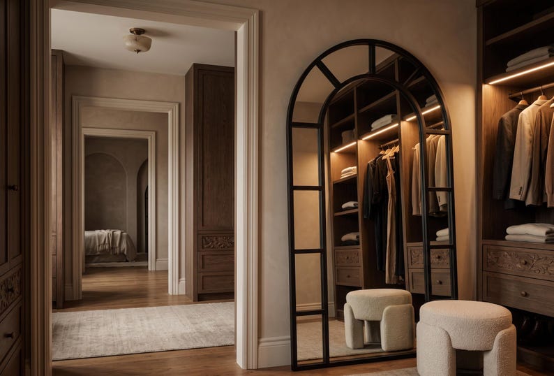 May include: A tall, arched floor mirror with a black metal frame featuring a grid design. The mirror reflects a walk-in closet with dark wood cabinetry, hanging garments, and integrated lighting. Two cream-colored, textured, cylindrical stools sit nearby.