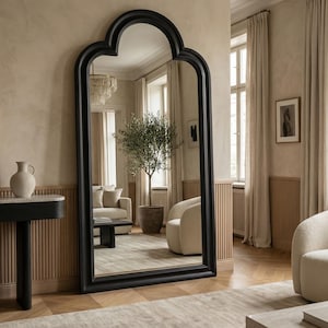 May include: A large, black-framed floor mirror with an arched top stands in a room with neutral tones. The mirror reflects a living space with a sofa, armchair, and a potted olive tree. The room features a console table and a patterned rug.