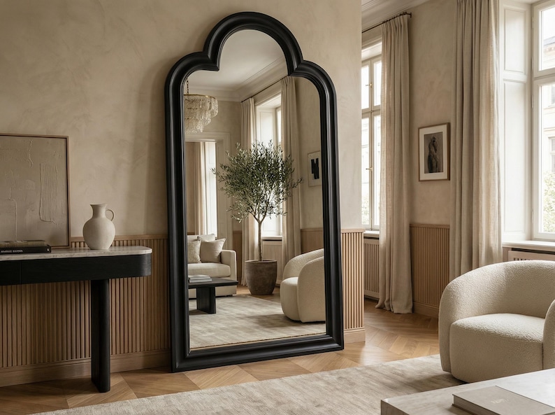 May include: A large, black-framed floor mirror with an arched top stands in a room with neutral tones. The mirror reflects a living space with a sofa, armchair, and a potted olive tree. The room features a console table and a patterned rug.