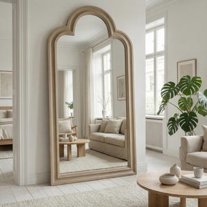 May include: A large, arched-top mirror with a light wood frame stands against a white wall. The mirror reflects a living room with a sofa, coffee table, and a large potted plant. The room has a neutral color palette.